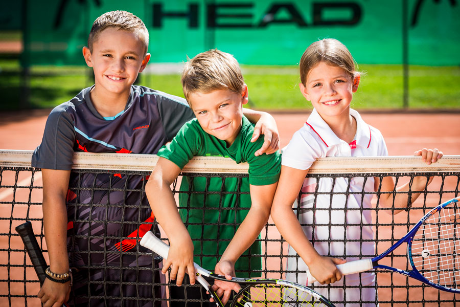 Kinder lehnen sich über Tennisnetz und halten ihre Schläger